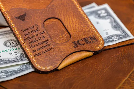 A handmade leather brown money clip wallet engraved with an inspirational quote and initials “JCEN,” holding U.S. dollar bills.