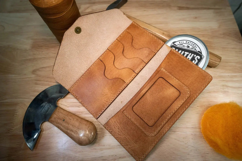 A premium leather long wallet, opened to show a spacious interior, with leather working tools surrounding it.