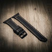 Black full-grain leather Apple Watch band with tan stitching and buckle on wood surface
