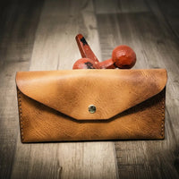 Handmade women's long wallet in saddle tan leather, featuring a brass button, on a wooden surface with a rustic red weight in the background.
