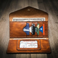 Women's leather wallet clutch in chestnut brown, handmade in the USA, displayed on a wooden surface with a rustic metal tool in the background.
