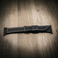 Apple Watch Leather Band in black full-grain leather with tan stitching on wood floor.